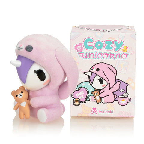 The official packaging for the Tokidoki Cozy Unicorno series, featuring the "Hug Me" theme and an illustration of Cuddle Bunny in a soft pink animal onesie.