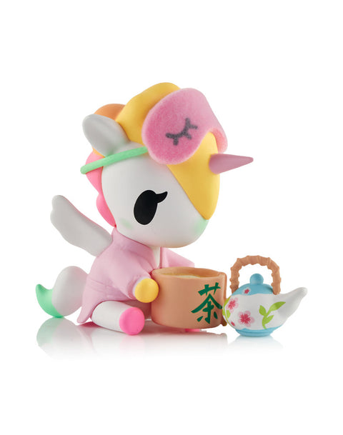 A white and yellow Unicorno figure wearing a pink sleep mask and robe, sitting next to a miniature white teapot with pink flowers and a cup of tea.