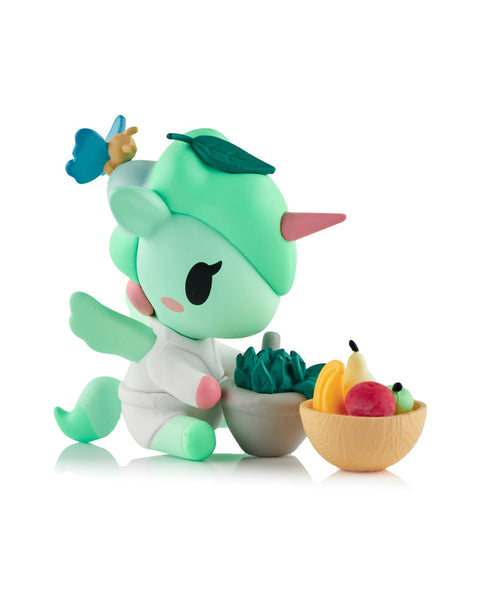 A pastel green Unicorno figure wearing a white spa robe, standing next to a grey mortar and pestle and a bowl filled with colorful fruit.