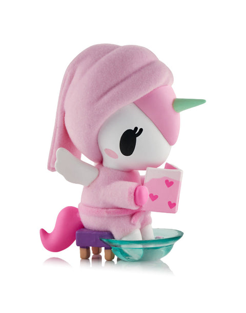 A white Unicorno figure wearing a pink towel head wrap, sitting on a purple stool with its hooves in a clear teal soaking bowl while reading a book.