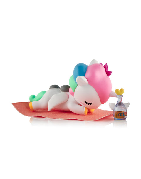 A white Unicorno figure with a pink and blue mane lying face down on an orange mat next to a small glass bottle of spa oil.