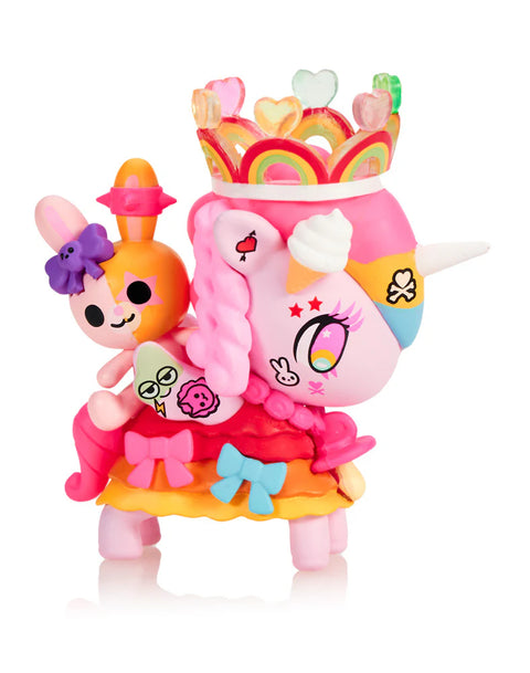 A bright pink Unicorno with braided hair wears a tall translucent heart crown with rainbow arches. A small orange rabbit sits on its back with expressive eye patches and printed symbols. The figure’s multicolored skirt features layered bows and textured ruffles, and star and heart markings decorate the face, giving a lively Harajuku look.