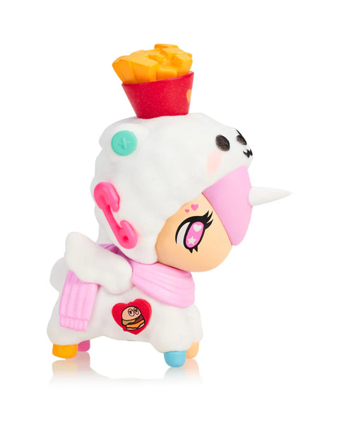 A white Unicorno figure designed like a soft fleece outfit has a pink scarf, pastel hooves, and a bright red container of yellow fries placed on its head. The figure’s face has pink accents, a small unicorn horn, and a heart patch featuring a burger. Buttons, patches, and molded textures enhance the playful food-themed design.