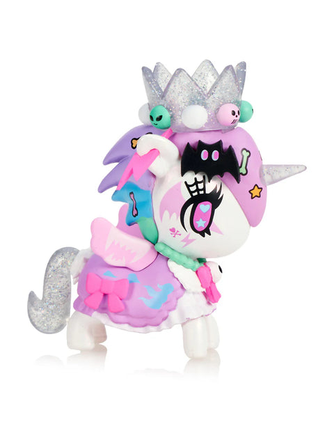 A pastel Unicorno decorated in creepy-cute style features a glittery silver crown topped with tiny skull and alien figures. Its lavender and aqua mane flows behind a pink bone accessory, and the black bat emblem on its face stands out. The figure wears a purple skirt with wing shapes and small bows, balancing eerie and sweet details.