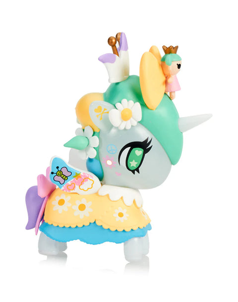 A soft gray Unicorno with green hair is decorated with daisy flowers, a butterfly wing pattern, and a small doll wearing a crown sitting behind its horn. Yellow and blue skirt layers show flower shapes, while pastel wings attach at the sides. The figure’s bright green eye and star markings emphasize its gentle, spring-inspired theme.