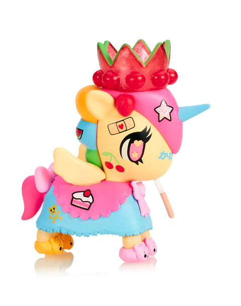 A yellow-and-pink Unicorno holds a small lollipop in its mouth and wears a red fruit-themed crown. Its skirt shows a slice of cake and skull-and-bones graphics, while the hooves resemble tiny slippers. The figure’s bright facial markings, layered skirt, and cheerful candy details emphasize a sweet, dessert-inspired theme.