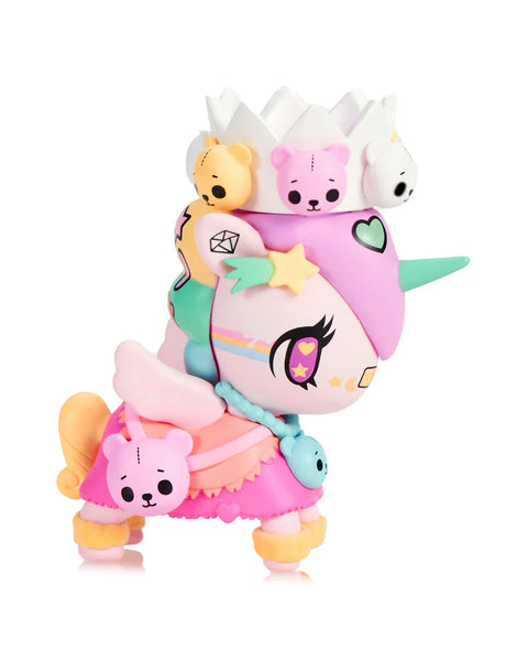 A pink-and-mint Unicorno wears a white crown surrounded by small teddy bear figures in pastel yellow, pink, and white. Additional teddy details rest on its skirt. The figure has soft star graphics, wing shapes, and textured braids. Heart-shaped patches and colorful accessories emphasize its plush-toy royal theme.