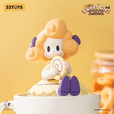 A pastel figurine girl with fluffy golden curls and purple bows, holding a yellow Swiss roll cookie. She wears a matching dress and sits atop a white cup, exuding soft bakery charm.