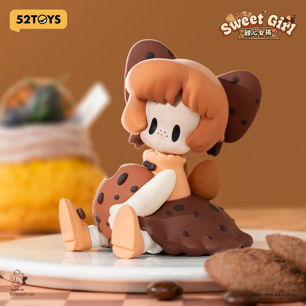 A whimsical figurine with chocolate brown bobbed hair, freckles, and oversized cookie-like ears. She wears a ruffled cookie skirt dotted like chocolate chips, sitting with a sweet shy expression.