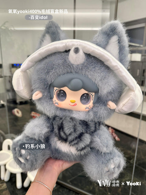 A plush wolf figure with gray fur and pointed ears is shown indoors. The doll has a serious yet cute face with small cheek tattoos, a white patch on its chest, and paw prints on its feet. It wears large, puffy ear muffs and embodies a stylish, cool idol persona.