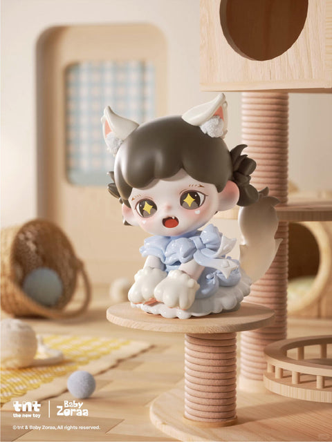 A vinyl figure with twin braids and white cat ears sits on a beige perch, smiling with bright yellow eyes. It wears a puffy blue outfit with bows and large paw-shaped feet, surrounded by yarn balls and wooden climbing posts in a sunlit cat playroom.