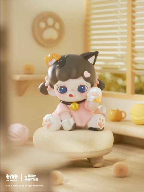A small cat-inspired figure with black ears and a pink outfit holds a milk bottle in one paw. It sits on a round beige cushion with yarn scattered around, wearing a bell collar and paw slippers. The soft golden light gives a warm, sleepy feeling.