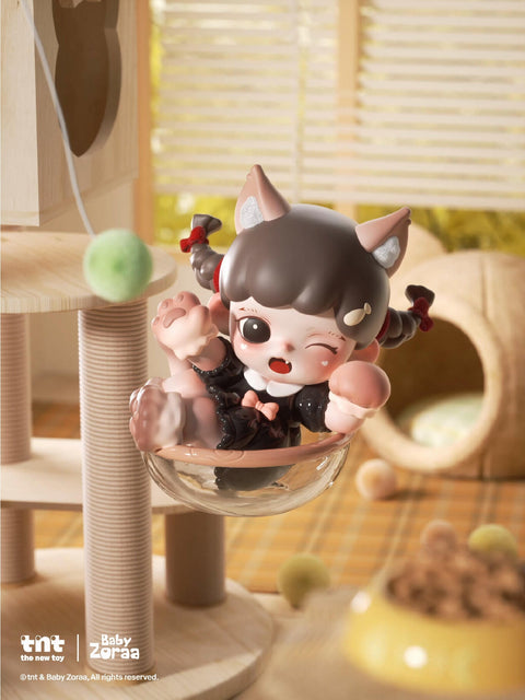 A small black-haired figure with cat ears sits in a transparent bowl chair, playfully kicking one paw while a green yarn ball swings nearby. The figure wears a black outfit with peach details and smiles with one eye closed.