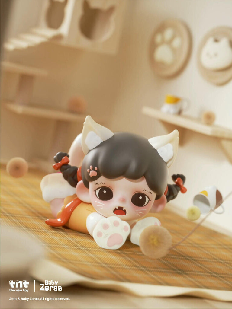 A tiny cat-girl figure with white ears lies belly-down on a checkered mat, reaching forward with both paws. Yarn balls and small props scatter around, showing a playful pose. Her expression is surprised, with fangs showing and pink paw pads visible.