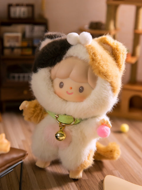 A close-up of the Clingy Calico Cat plush figure from the Finding Unicorn x 卓大王 “My Little Cat Series” blind box collection. The doll features a soft, smiling face with gray hair, dressed in a fluffy white costume with calico patches of orange, black, and brown. It wears a mint green collar with a shiny gold bell and holds a small pink pom-pom. The background is a cozy, cat-themed mini room with warm lighting and wooden flooring.