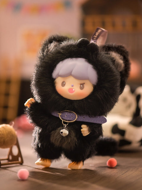 A close-up of the Naughty Black Cat plush from the Finding Unicorn x 卓大王 “My Little Cat Series” blind box collection. The doll has lavender hair, a mischievous expression with a tiny fang showing, and wears a fluffy black cat costume with pointed ears and a purple collar featuring a silver bell. Set in a warm, cozy mini room with wooden flooring and scattered pom-poms, the scene captures a playful and charming cat-inspired atmosphere.