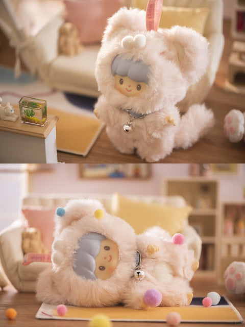Two cozy scenes featuring the Sweet Pink Tabby Cat plush from the Finding Unicorn x 卓大王 “My Little Cat Series” blind box. The doll has a gentle smile, light blue hair, and wears a fluffy pastel pink cat costume with a white belly, soft ears, and a light blue collar with a silver bell. One image shows the plush standing in a warmly lit living room; the other shows it lying on a rug surrounded by colorful pom-poms, evoking a playful and comforting atmosphere.
